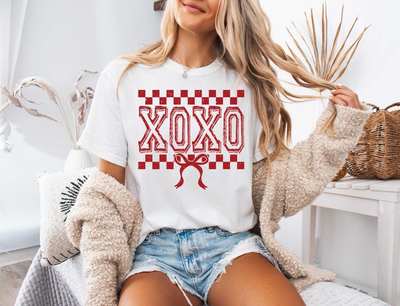 以下が含まれることがあります： 赤いチェック柄の白いTシャツに、赤い「XOXO」の文字と、その下に赤いリボンが描かれています。