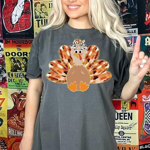 May include: A dark gray t-shirt with a graphic of a turkey wearing a hat. The turkey is brown with orange and brown feathers. The turkey is surrounded by a wreath of orange and brown leaves.