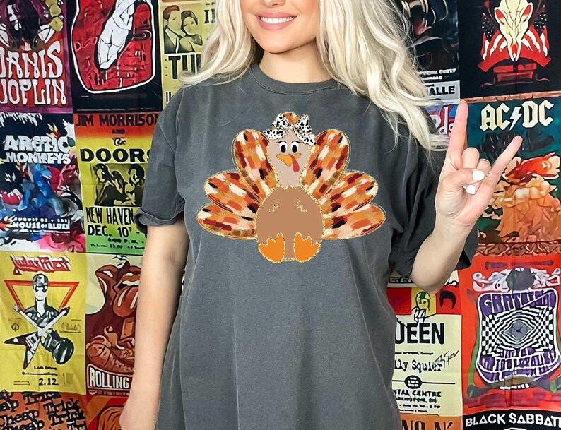 May include: A dark gray t-shirt with a graphic of a turkey wearing a hat. The turkey is brown with orange and brown feathers. The turkey is surrounded by a wreath of orange and brown leaves.