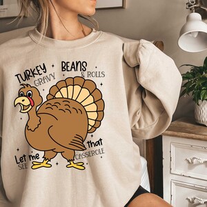 May include: Beige sweatshirt with a Thanksgiving-themed graphic. The graphic features a cartoon turkey with the text "Let me see that casserole" and "Turkey Gravy Beans & Rolls".