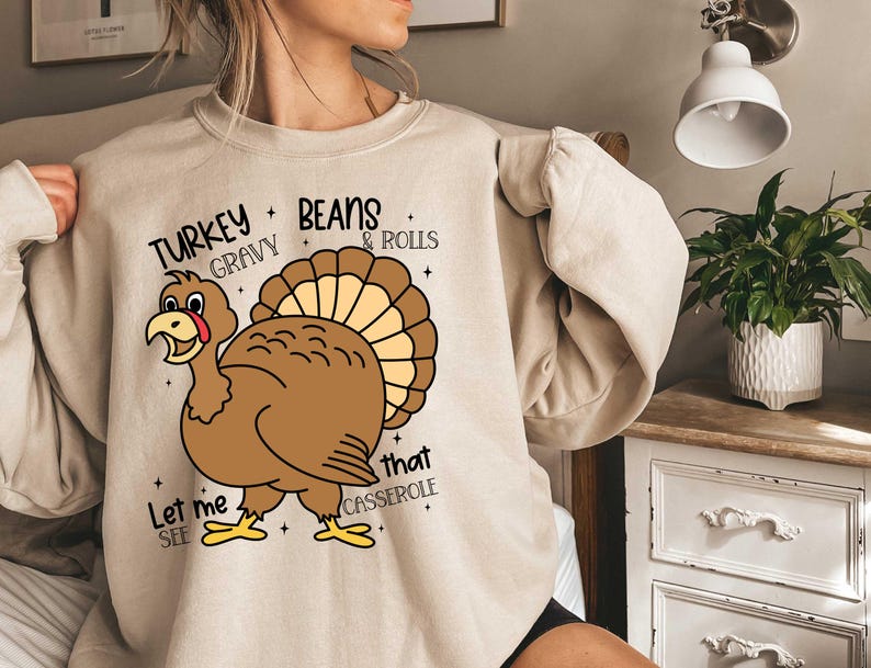 May include: Beige sweatshirt with a Thanksgiving-themed graphic. The graphic features a cartoon turkey with the text "Let me see that casserole" and "Turkey Gravy Beans & Rolls".