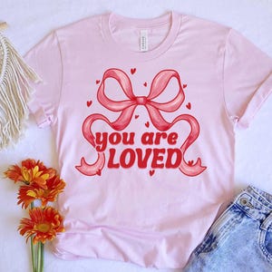 以下が含まれることがあります： 赤いリボンと「you are loved」というレトロなフォントのテキストが書かれたピンクのTシャツです。