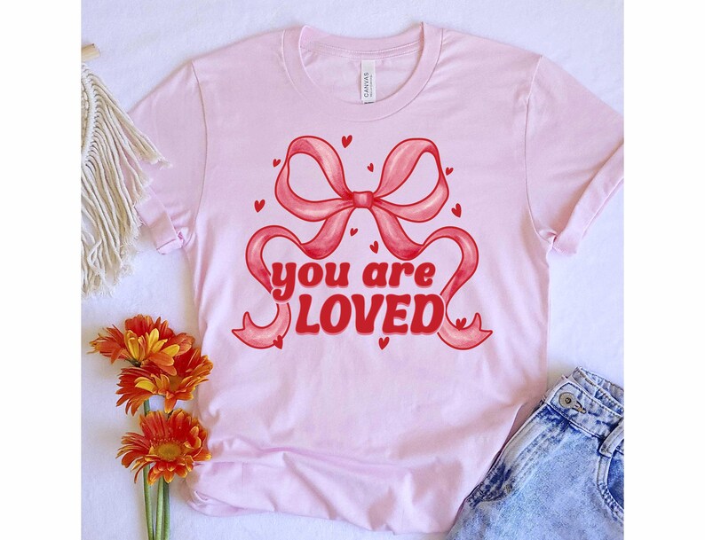 以下が含まれることがあります： 赤いリボンと「you are loved」というレトロなフォントのテキストが書かれたピンクのTシャツです。