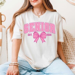 May include: Cream-colored t-shirt with the word "SENIOR" in pink block letters, along with "EST. 2026" and a pink bow graphic. The shirt has short sleeves and a relaxed fit.