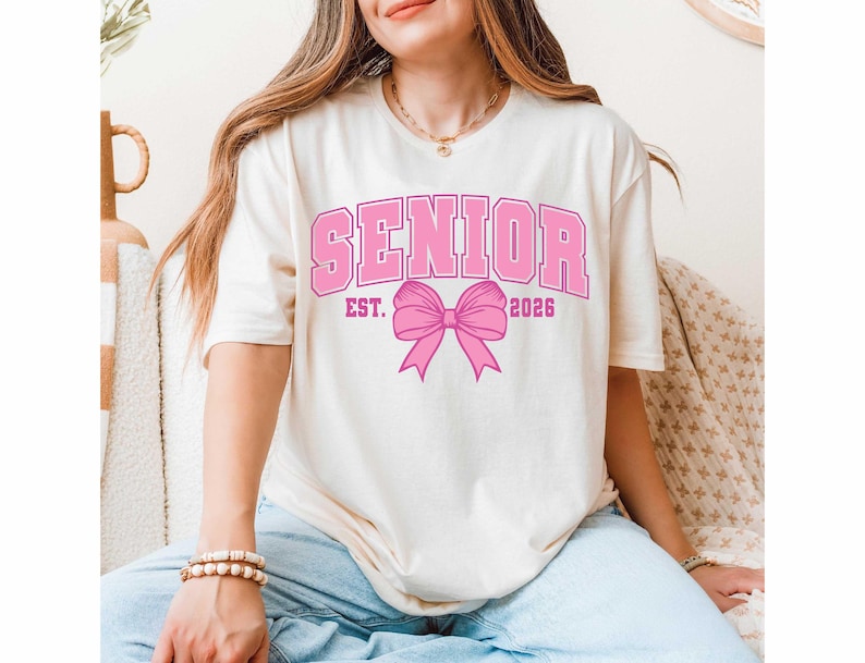 May include: Cream-colored t-shirt with the word "SENIOR" in pink block letters, along with "EST. 2026" and a pink bow graphic. The shirt has short sleeves and a relaxed fit.