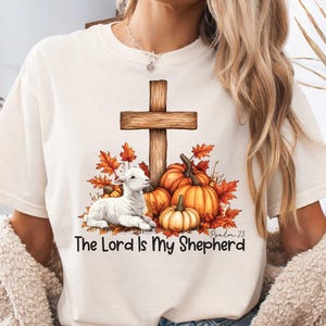 May include: White t-shirt with a brown wooden cross, a white lamb, and fall-colored leaves and pumpkins. The text "The Lord is My Shepherd" and "Psalm 23" are printed on the shirt.