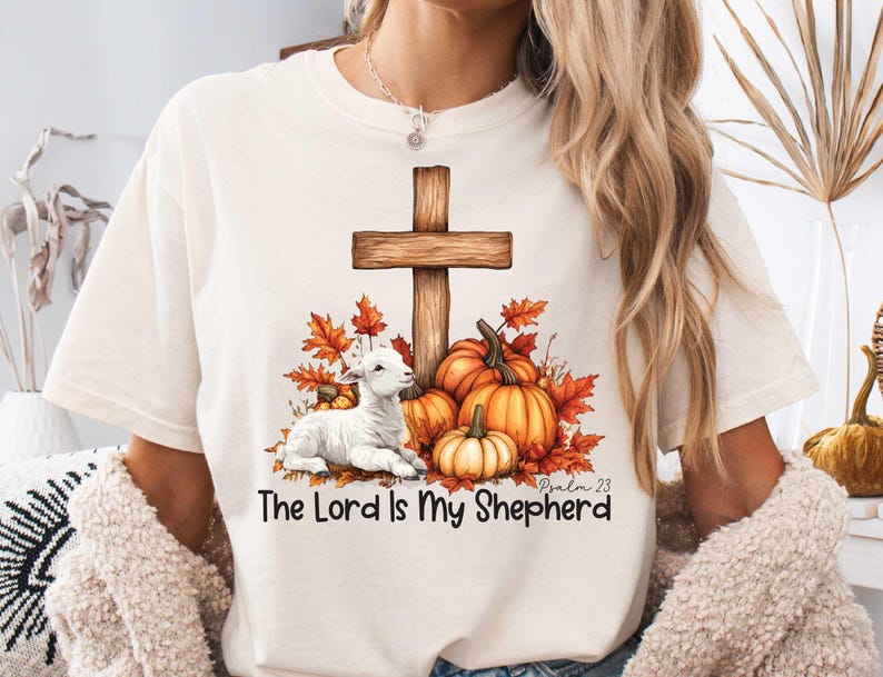 May include: White t-shirt with a brown wooden cross, a white lamb, and fall-colored leaves and pumpkins. The text "The Lord is My Shepherd" and "Psalm 23" are printed on the shirt.