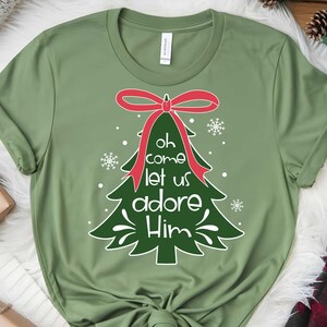 May include: An olive green t-shirt with a Christmas tree design. The tree has a red bow and the words "Oh come let us adore Him" in white. The design includes snowflakes. The shirt is made of a soft material.