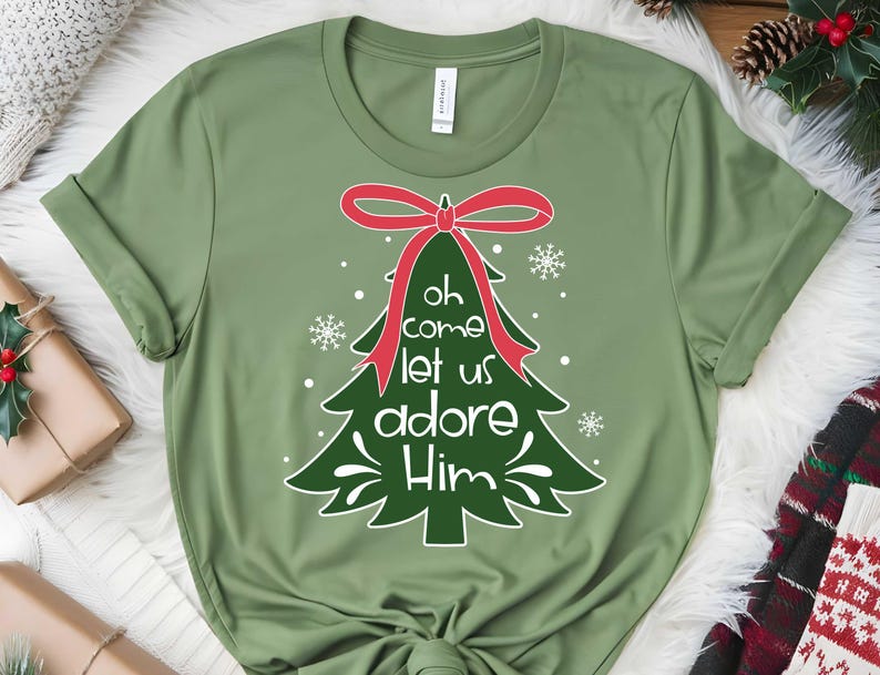 May include: An olive green t-shirt with a Christmas tree design. The tree has a red bow and the words "Oh come let us adore Him" in white. The design includes snowflakes. The shirt is made of a soft material.