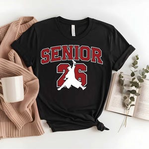 May include: Black t-shirt with the text "SENIOR 26" in red and white. The design includes a silhouette of a person in a graduation gown. A beige sweater, a white mug, and an open book are also visible.