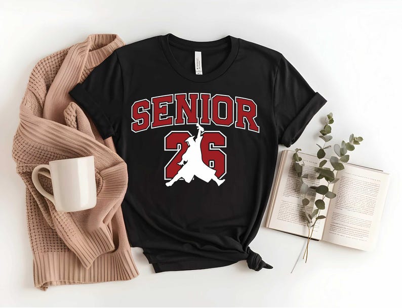 May include: Black t-shirt with the text "SENIOR 26" in red and white. The design includes a silhouette of a person in a graduation gown. A beige sweater, a white mug, and an open book are also visible.