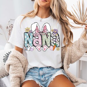 May include: White t-shirt with a colorful Easter-themed graphic that says "Nana" with bunny ears, flowers, and eggs.