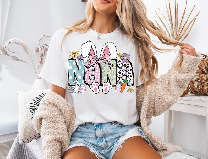 May include: White t-shirt with a colorful Easter-themed graphic that says "Nana" with bunny ears, flowers, and eggs.