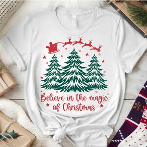 May include: White t-shirt with a Christmas design. The graphic features green Christmas trees, red stars, and Santa Claus in a sleigh pulled by reindeer. The text reads "Believe in the magic of Christmas."