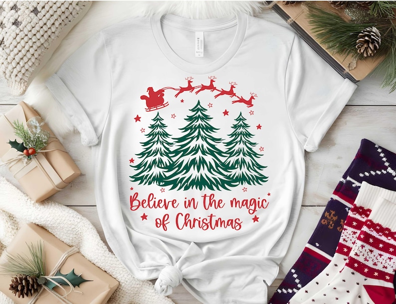 May include: White t-shirt with a Christmas design. The graphic features green Christmas trees, red stars, and Santa Claus in a sleigh pulled by reindeer. The text reads "Believe in the magic of Christmas."