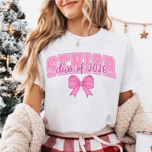 May include: White t-shirt with the text "SENIOR class of 2026" in pink block letters and a pink bow graphic. The shirt is a casual style, suitable for graduation celebrations.
