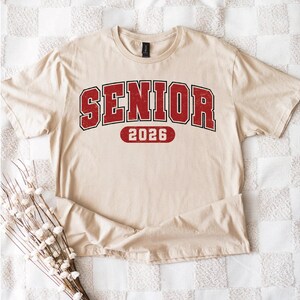 May include: A beige t-shirt with the word "SENIOR" in red, outlined in black, and the year "2026" in red. The shirt is folded on a checkered surface, with a bundle of white flowers in the lower left corner.