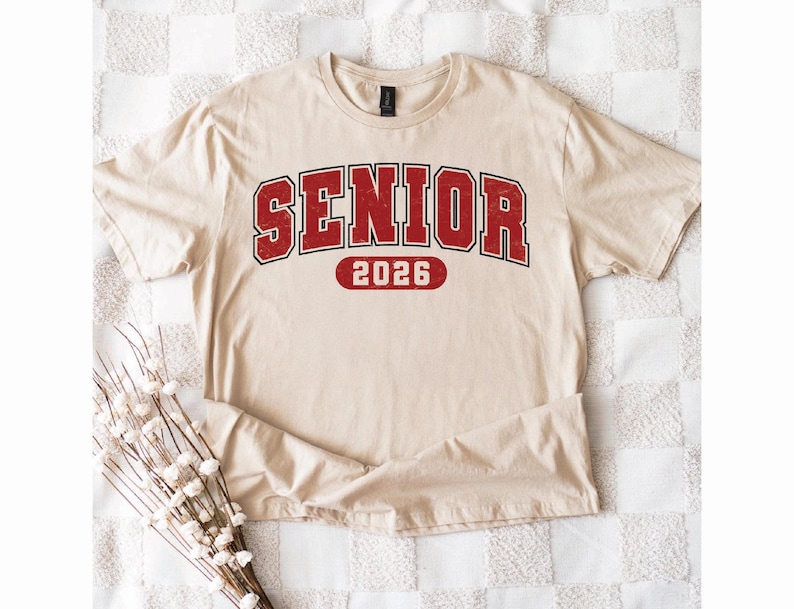 May include: A beige t-shirt with the word "SENIOR" in red, outlined in black, and the year "2026" in red. The shirt is folded on a checkered surface, with a bundle of white flowers in the lower left corner.