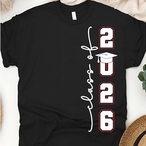 May include: Black t-shirt with the words "Class of 2026" in white script and red-outlined numbers. A graduation cap graphic is above the "2026" text. The shirt is a classic crew neck style.