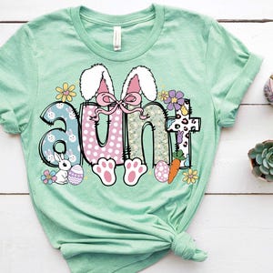 May include: A light green t-shirt with a graphic of the word "aunt" in colorful letters with a bunny rabbit design. The letters are decorated with flowers, polka dots, and a carrot.