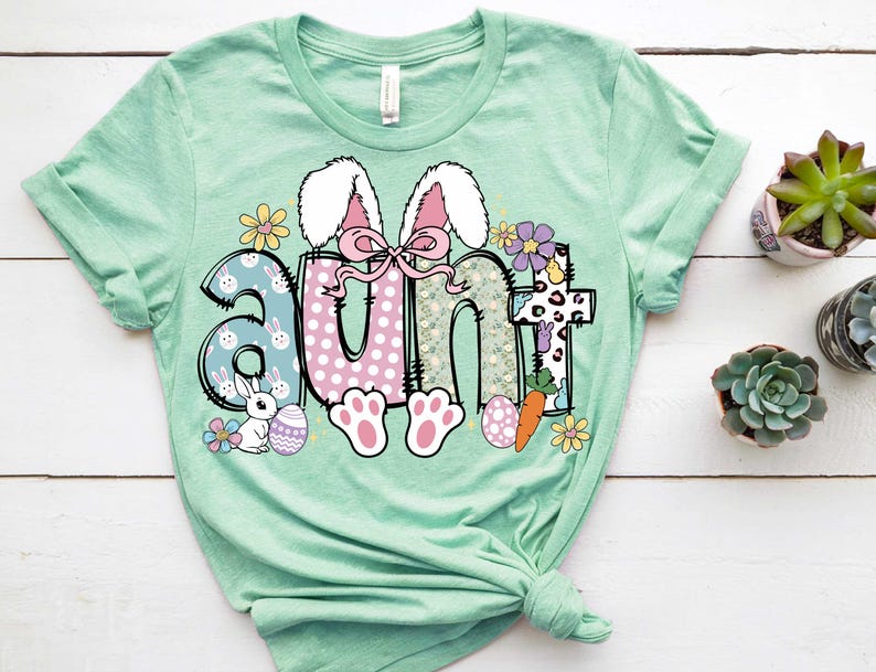 May include: A light green t-shirt with a graphic of the word "aunt" in colorful letters with a bunny rabbit design. The letters are decorated with flowers, polka dots, and a carrot.