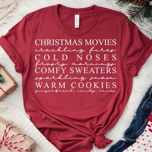 May include: A crimson t-shirt featuring white text with Christmas-themed words: Christmas movies, crackling fires, cold noses, frosty mornings, comfy sweaters, sparkling snow, and warm cookies. The shirt is knotted at the hem.