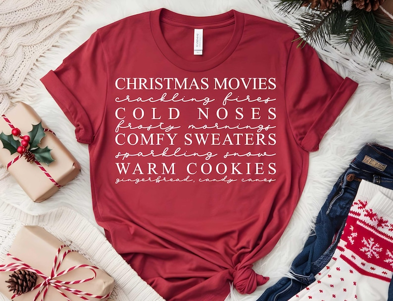 May include: A crimson t-shirt featuring white text with Christmas-themed words: Christmas movies, crackling fires, cold noses, frosty mornings, comfy sweaters, sparkling snow, and warm cookies. The shirt is knotted at the hem.