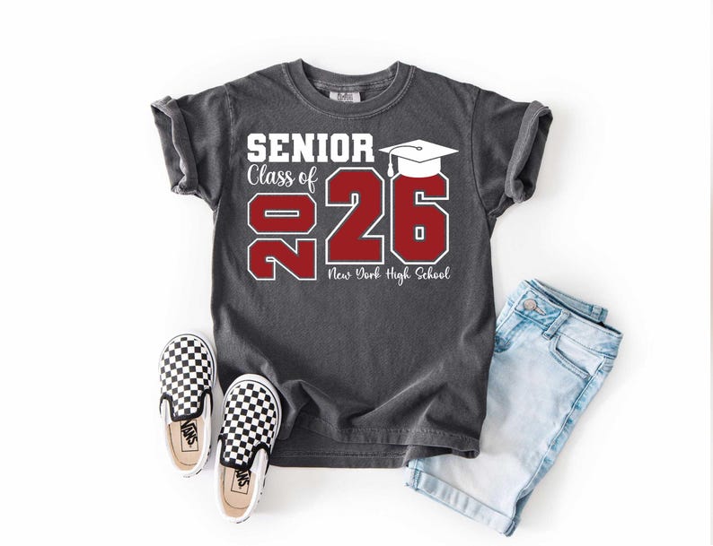 May include: Dark gray t-shirt with the text "SENIOR Class of 2026" in white and red. The shirt features a graduation cap graphic and the words "New York High School". The shirt is paired with light blue denim shorts and black and white checkered sneakers.