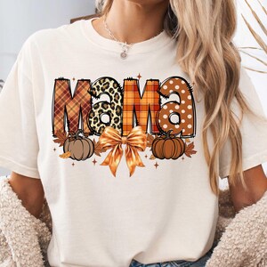 May include: White t-shirt with the word "Mama" in orange, brown, and black patterned letters. The letters are decorated with pumpkins, leaves, and a bow.