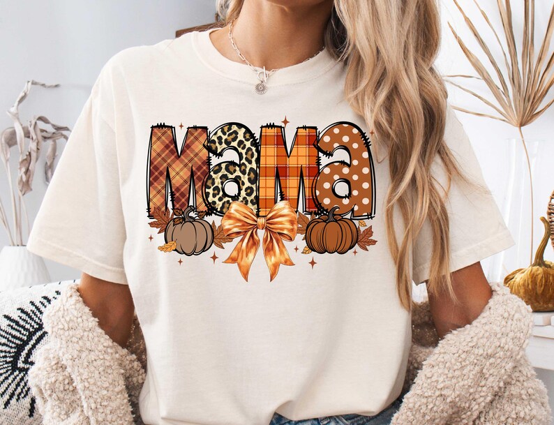 May include: White t-shirt with the word "Mama" in orange, brown, and black patterned letters. The letters are decorated with pumpkins, leaves, and a bow.