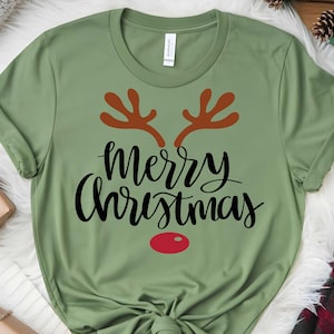 May include: A green t-shirt featuring the words "Merry Christmas" in black script, brown antlers, and a red nose. This festive t-shirt is a holiday apparel item.