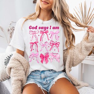 以下が含まれることがあります： ピンクの文字で「God says I am」と書かれた白いTシャツ。テキストは、「愛されている」「強い」「価値がある」「重要」などの言葉が書かれたピンクのリボンで囲まれています。