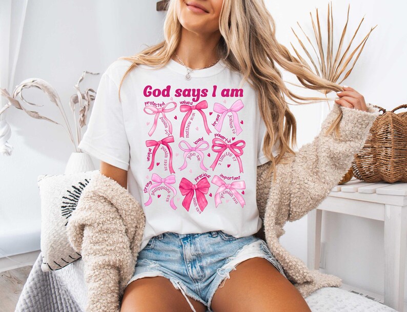 以下が含まれることがあります： ピンクの文字で「God says I am」と書かれた白いTシャツ。テキストは、「愛されている」「強い」「価値がある」「重要」などの言葉が書かれたピンクのリボンで囲まれています。