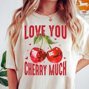 以下が含まれることがあります： 「Love You Cherry Much」という赤い文字が書かれた白いTシャツ。文字はかすれたフォントで書かれています。文字の下には、ディスコボールのデザインが施された赤いチェリーが2つ描かれています。