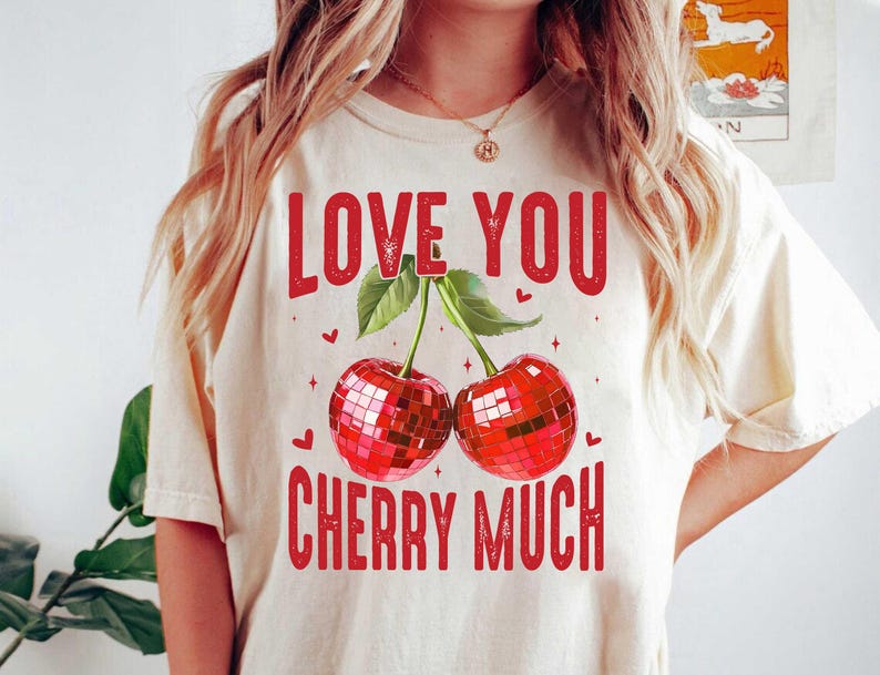 以下が含まれることがあります： 「Love You Cherry Much」という赤い文字が書かれた白いTシャツ。文字はかすれたフォントで書かれています。文字の下には、ディスコボールのデザインが施された赤いチェリーが2つ描かれています。