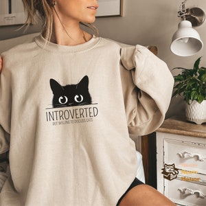 May include: A beige sweatshirt with a black cat peeking over the top and the text "Introverted but willing to discuss cats".