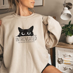 Könnte beinhalten: Ein beiger Pullover mit einer schwarzen Katze, die über den Rand schaut, und dem Text "Introvertiert, aber bereit, über Katzen zu diskutieren".