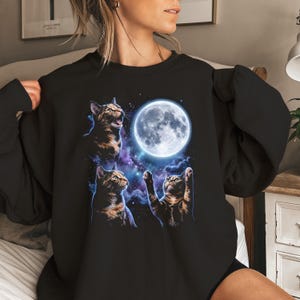 Può includere: Una felpa nera a maniche lunghe con una stampa grafica di tre gatti tigrati che guardano verso l'alto una luna piena luminosa e brillante su uno sfondo cosmico scuro, viola e blu, con stelle bianche sparse e luce celestiale.
