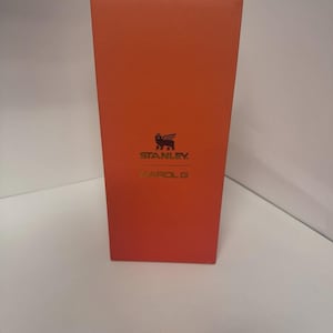 May include: A rectangular orange box featuring the black Stanley logo with a winged bear icon at the top and the text KAROL G printed in gold lettering below the logo. The packaging is oriented vertically with a smooth, solid orange finish.