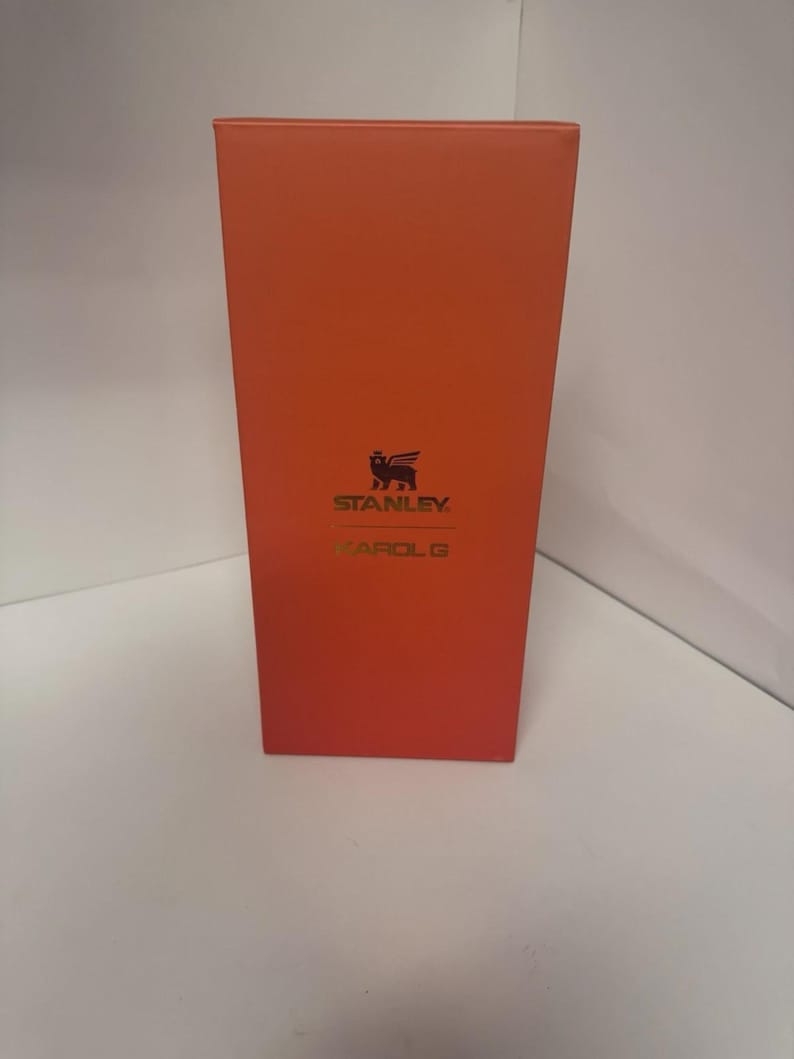 May include: A rectangular orange box featuring the black Stanley logo with a winged bear icon at the top and the text KAROL G printed in gold lettering below the logo. The packaging is oriented vertically with a smooth, solid orange finish.