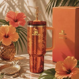 May include: A metallic copper-toned Stanley tumbler with a floral pattern, a handle, and a gold-colored pineapple-shaped straw topper. Beside it stands a matching orange box labeled Stanley KAHLO G, with hibiscus flowers and a pineapple dish nearby.