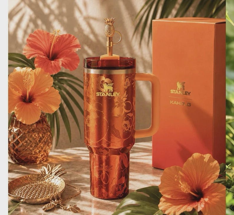May include: A metallic copper-toned Stanley tumbler with a floral pattern, a handle, and a gold-colored pineapple-shaped straw topper. Beside it stands a matching orange box labeled Stanley KAHLO G, with hibiscus flowers and a pineapple dish nearby.