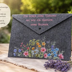 Könnte beinhalten: Eine graue Filz-Umschlagtasche mit floralem Design und dem Text "EIN LEBEN OHNE TRÄUME IST WIE EIN GARTEN OHNE BLUMEN." Die Tasche hat einen Knopfverschluss und ist mit bunten Blumen verziert.