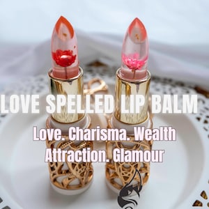 Könnte beinhalten: Zwei goldene und weiße Lippenbalsame mit durchsichtigen Röhrchen, die eine rote und eine rosa Blume enthalten. Die Lippenbalsame haben den Text "Love. Charisma. Wealth Attraction. Glamour" auf der Vorderseite aufgedruckt.