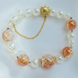 May include: A white pearl bracelet with a gold sun charm and orange glass beads. The bracelet has a delicate gold chain attached to the sun charm.