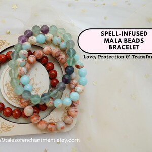 Puede incluir: Un conjunto de cuatro pulseras de cuentas mala de colores, apiladas sobre una superficie de madera. Las pulseras están hechas de cuentas rojas, azules, verdes y blancas. El texto "SPELL-INFUSED MALA BEADS BRACELET Love, Protection & Transformation" está escrito en una pancarta rosa.
