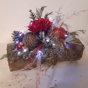 Peut inclure: Une décoration de Noël festive sur une base en bois, comprenant une fleur rouge, des pommes de pin et des ornements décoratifs. L'arrangement est orné de guirlandes lumineuses scintillantes, créant une ambiance chaleureuse.