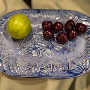 May include: An oval ceramic serving platter with a blue and white glazed finish featuring a repeating geometric star pattern. A single bright green lime and six dark red cherries are arranged on the textured surface of the dish.