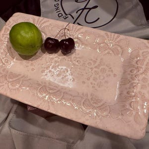 May include: A rectangular light pink ceramic tray with a raised, intricate lace-like floral pattern across its surface. A single bright green lime and two dark red cherries are placed on the left side of the tray, contrasting with the pale pink glaze.