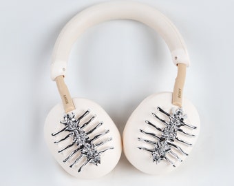 May include: White over-ear headphones with a beige headband and silver accents on the earcups. The earcups have a unique design with a silver skeletal pattern.